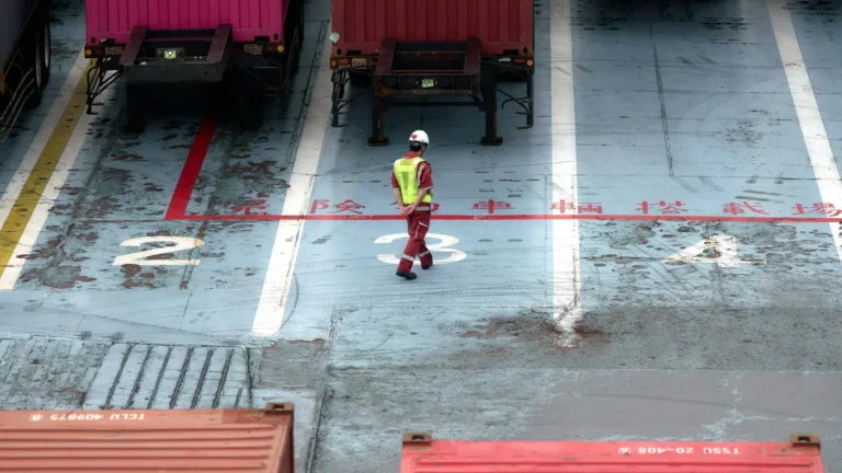 Worker in a hi-vis vest and hard hat walking past container trailers parked in numbered bays
