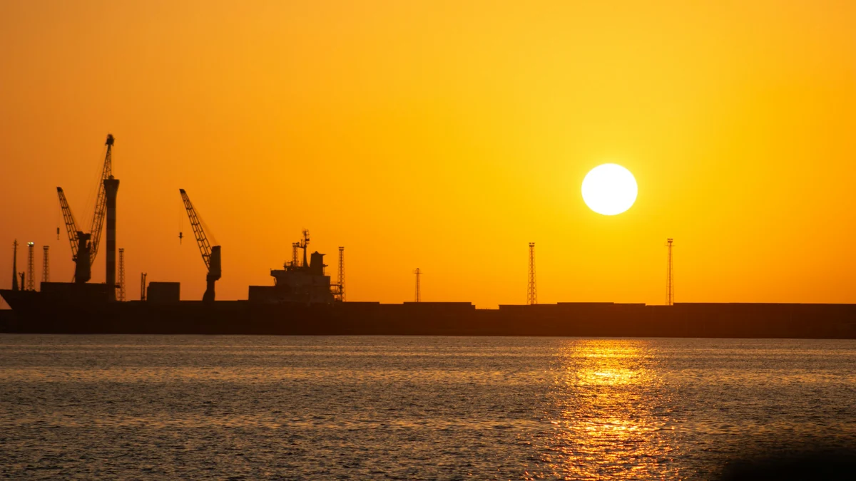 Silhouettes of harbor cranes and a moored cargo ship against an orange sunset over calm water