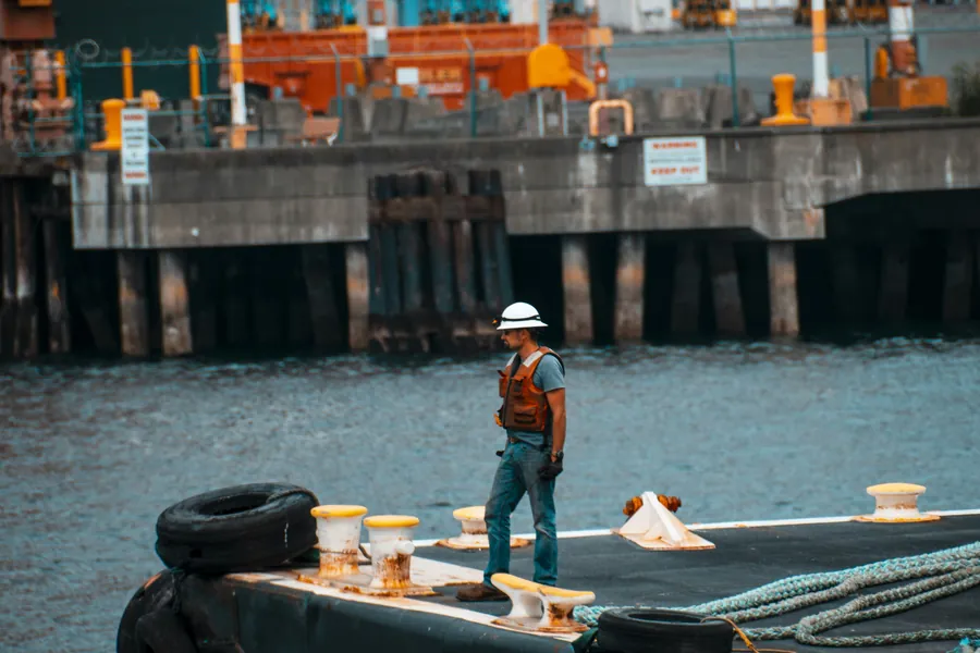 Worker in a hard hat and orange vest standing on a moored barge deck beside a harbor pier
