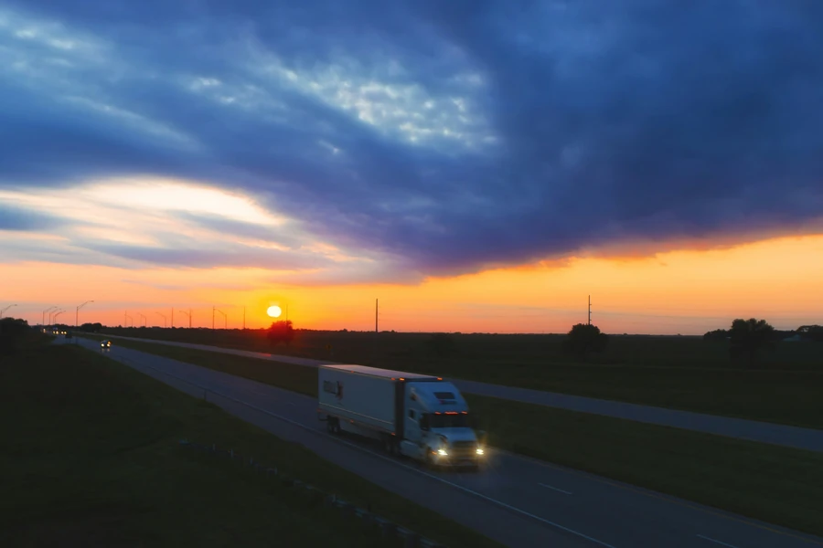 Semi-truck with headlights on driving along an empty highway at sunset under dark blue clouds