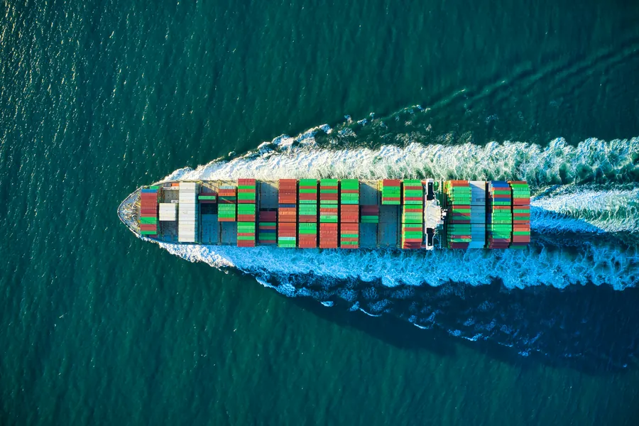 Aerial view of a container ship stacked with colorful containers cutting a white wake through green sea