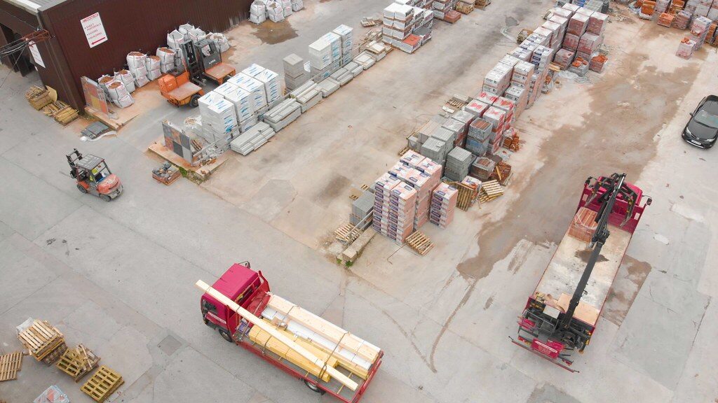 Aerial view of a freight yard with a flatbed truck hauling strapped timber and a crane truck loading pallets
